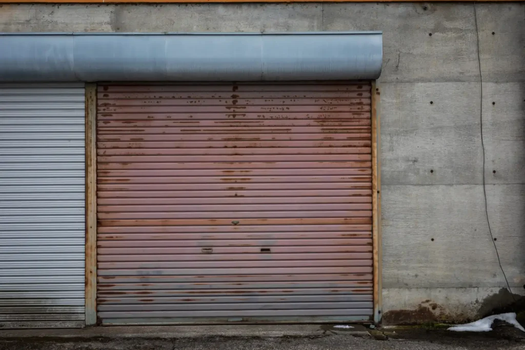 A closed, rusty metal garage door stands against a concrete wall, showing signs of wear beneath a slight overhang above.