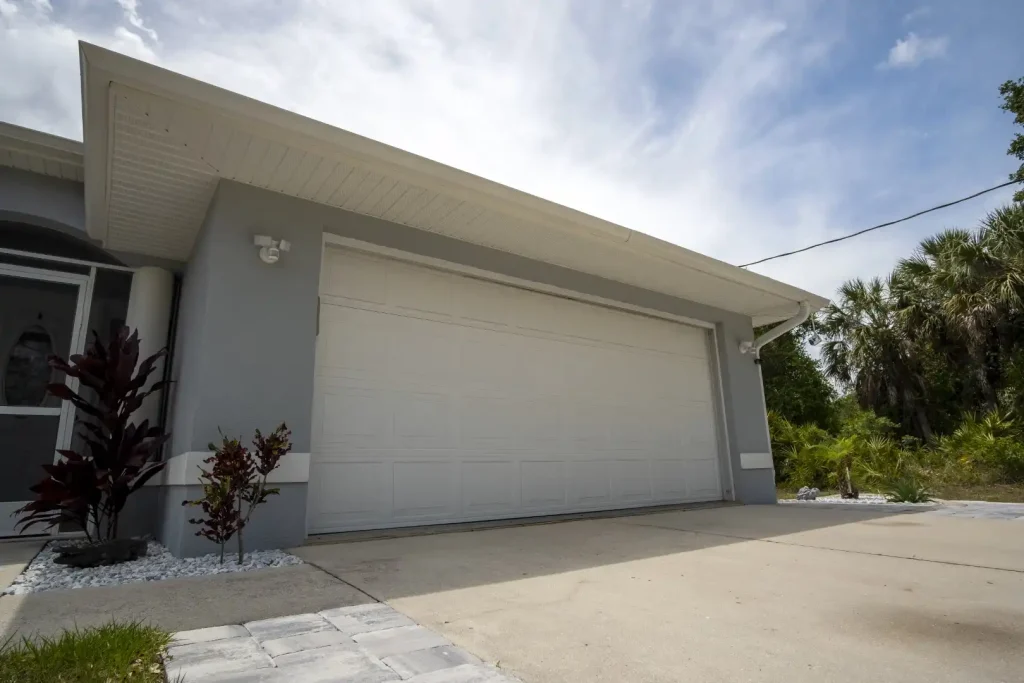 A pristine white garage door stands against the sleek gray building, complemented by lush plants and a concrete driveway. Under a cloudy sky, it's an upgrade that enhances curb appeal.