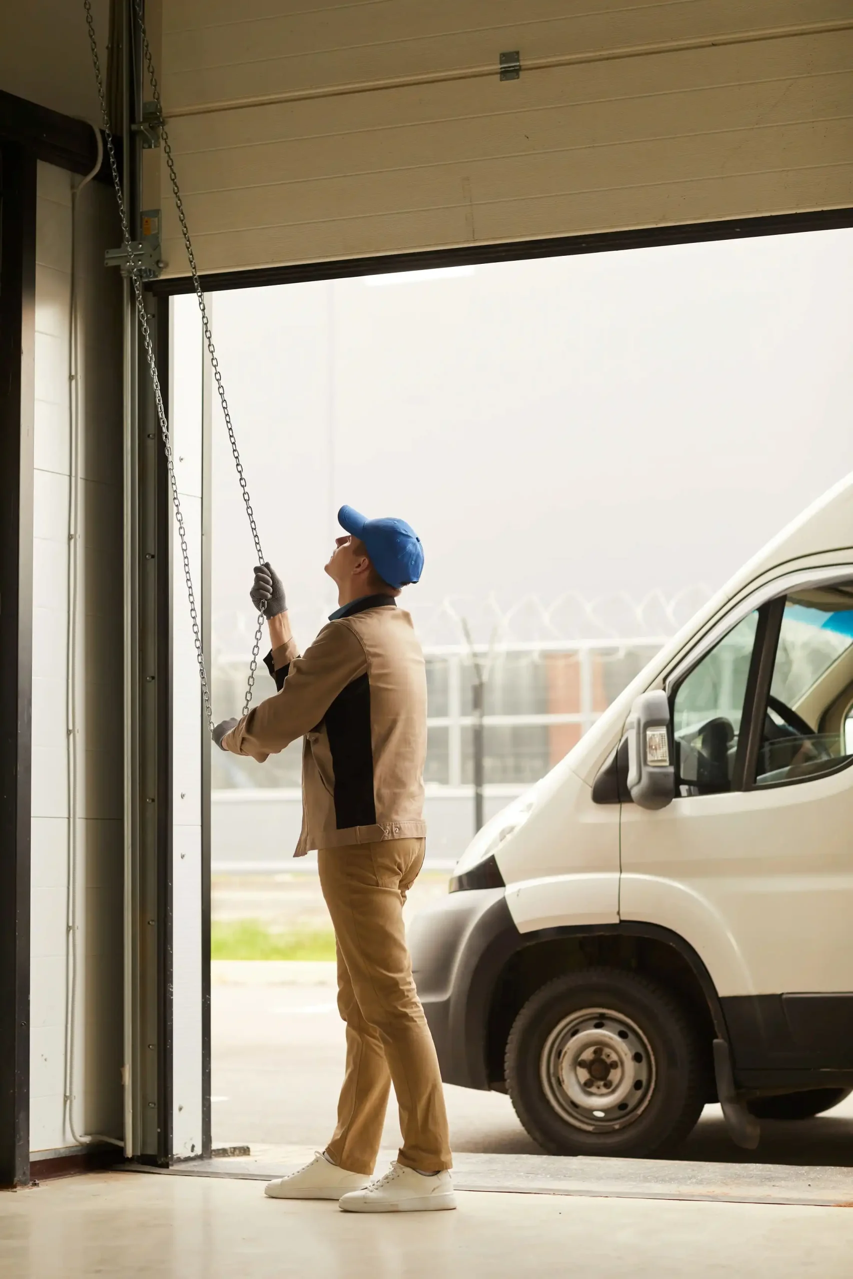 In work attire, a person pulls a chain to open the garage door, following essential maintenance tips to extend its life. A white van is parked outside.