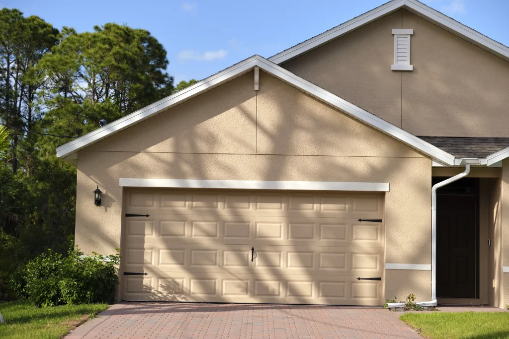 Beige house with a two-car garage door and single outdoor light fixture, nestled amid trees under a clear blue sky. Regular maintenance tips can help extend the life of your garage door, adding durability to this picturesque abode.