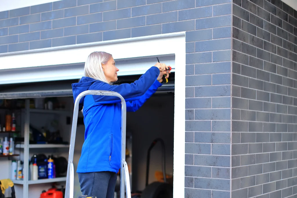 Person in a blue jacket on a ladder uses a screwdriver at the garage entrance, offering essential maintenance tips for ensuring your garage door operates smoothly.